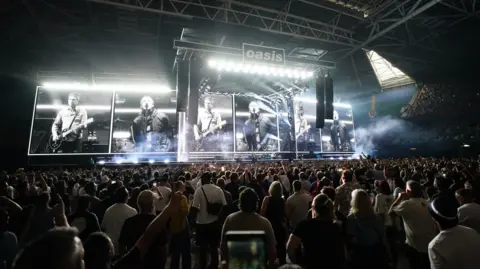 PA Media Crowds at the Oasis concert standing and facing the stage. A screen behind the stage shows the Gallagher brothers in black and white, Noel playing a guitar and Liam singing. The Oasis band logo can be seen at the top of the stage. 