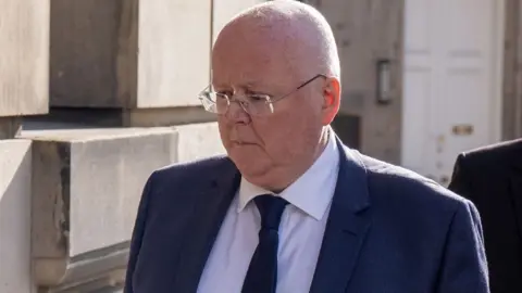 Peter Murrell, who is bald with glasses, looks downwards as he walks past a stone building 