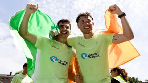 Two men are standing side-by-side, holding up an Irish tricolour flag - which is three vertical stripes of green, white and orange - are posing for photographs. They are standing with their arms outstretched, almost around each other, as they hold out the flag behind their backs. They are both wearing light-green running t-shirts, and have short dark hair and broad smiles. In the background two people are visible looking towards one another. 