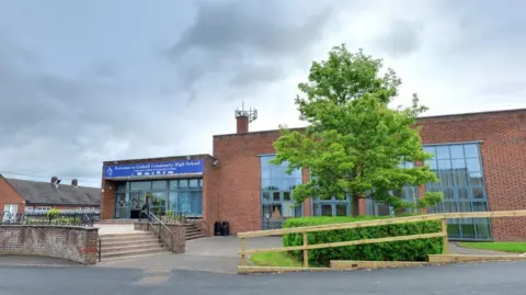 The entrance to Codsall Community High School with steps up to a set of glass doors. There is a large tree in front of a building with floor to ceiling windows. There is a blue sign above the entrance.