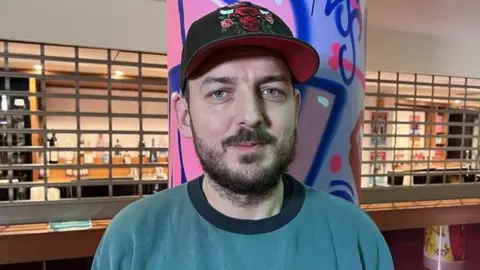 Kris Holland/BBC Jesse Jones who is wearing a blue jumper and a Chicago Bulls baseball cap. He has dark hair and a beard and is standing in the foyer of the Derngate theatre.
