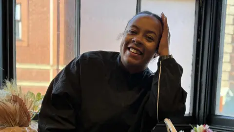 Pia Carson-Moore Pia Carson-Moore smiles at the camera with one of her hands resting on her head. She has dark hair that has been tied up behind her head and she wears a black sports jacket. She is sitting in front of a large window.