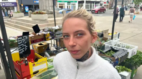 Olga Malolepsza, has blonde hair pulled back over her head, she has a butterfly tattoo on her neck under a zipped up white woollen jumper, behind her is her fruit and veg stall with cars and pedestrians passing.