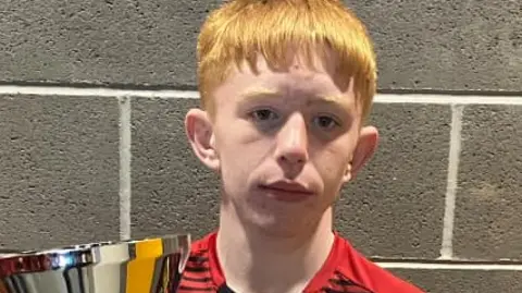 Kory McCrimmon who has ginger hair. He is looking at the camera and is wearing a red T-shirt