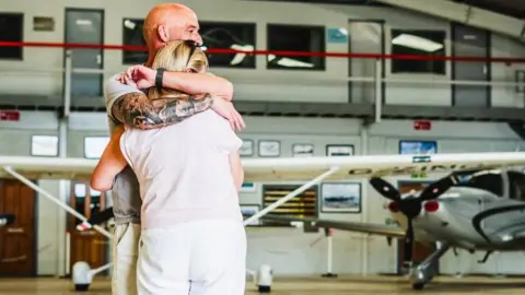 Elite Competitions A man and woman are seen hugging. There is a plane in the background, on the right of the photo.