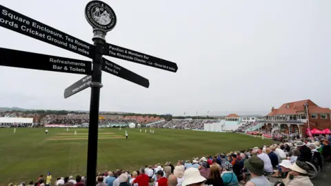 Getty Images A crowd of people sit in stands around a cricket pitch as a match takes place.