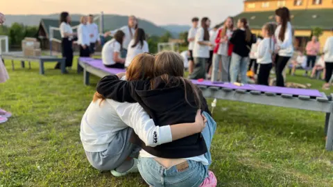 BBC Two teenage girls sit with their arms around each other, hugging, while sitting in front of a camp fire