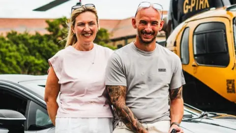 Elite Competitions A man and woman are seen sat on a car. The woman has a pale pink top and blonde hair with sunglasses on her head. The man has a grey top, tattoos on his arms and sunglasses on his head. They are both sat in front of a yellow helicopter