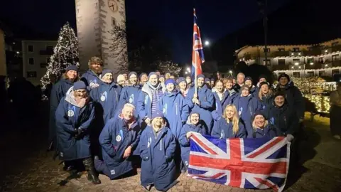 The International Ice Swimming Association (IISA) GB team posing for a picture with the union flag. One contestant is also holding a flag on a pole. There is a Christmas tree behind them. It's night time.