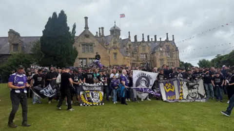 A large group of Erzgebirge Aue fans holding flags and banners standing outside Brownlow House - a brown brick building in a Baronial style