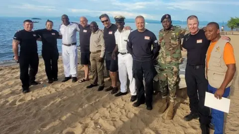 London Fire Brigade A line of 11 people including six firefighters, wearing their black uniform, stood alongside five Malawian delegates from the Defence Force, police, Port Authority and Department of Fisheries, all on the beach smiling at the camera.  