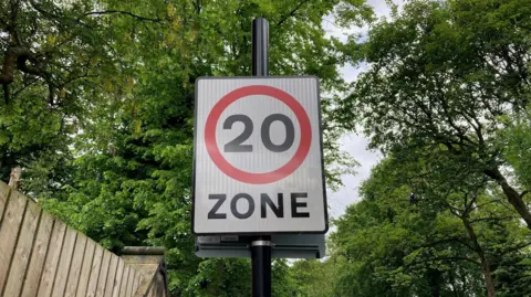 Close-up of a road sign showing 20 miles per hour in black writing surrounded by a red circle with a white background.