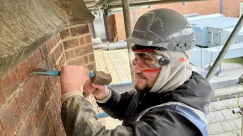 A man in a black dusty jacket with a grey helmet and eye goggles chiselling at a brick wall. 