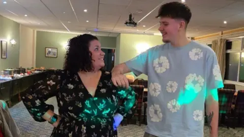Deniz Mills A woman stands next to a taller teenager in a function room, both looking at each other. She has black curly bobbed hair and is wearing a black wrap dress with flowers on it and he has dark brown hair and a goatee and is wearing a sky blue t-shirt with white flowers on it. Behind them are tables with silver pots of hot food, long tables and covered chairs.