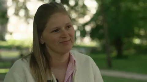 Charlotte, who has long light-brown hair and is wearing a white cardigan and pink and white striped shirt, is sitting in a park 