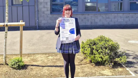Bedford Giving Elizabeth, a girl with bright red hair, glasses, and a skirt on with tights, and a blazer, holding up a certificate, by a school, with a bush and trees around her. 