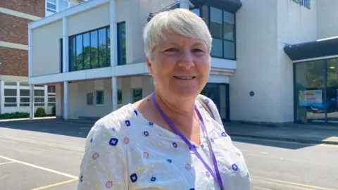 Jill Barker is smiling at the camera and is standing outside Strode Theatre. She is wearing a white blouse.