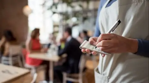 A close-up shot of a person's hands on a notepad taking an order, with a silver and black pen. They are wearing a cream top with pockets and blue sleeves. In the blurred background there are people sat at tables having dinner. 