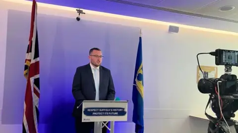 Vikki Irwin/BBC Michael Hadwen wearing a dark suit, white shirt and light blue tie. He is standing behind a lectern which says "RESPECT SUFFOLK'S HISTORY PROTECT ITS FUTURE" on its front. The union jack and the Suffolk county flags are hanging down on either side of him.