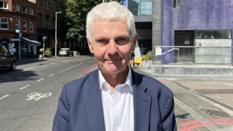 Grey-haired man in white shirt and navy jacket smiles at camera