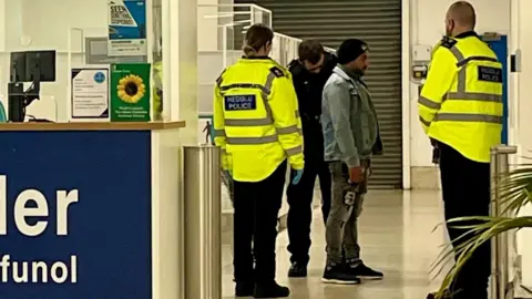 A man being detained at the UK Border by police. There are two officers in hi-vis and black trousers and another officer in all black. The main being detained has a black woolly hat on, a denim jacket and denim jeans. 