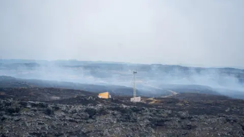 TERRES AUSTRALES ET ANTARCTIQUES FRANCAISES An expanse of burnt fields with smoke rising into the air. A radio mast and a small hut stands at the centre of the frame.