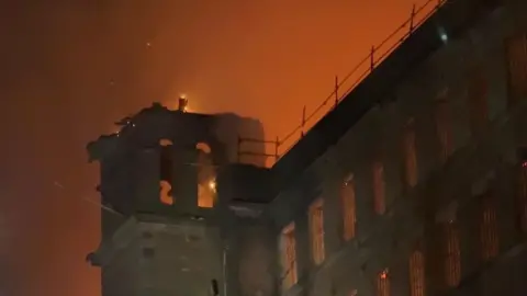 The upper levels of a brick mill building that is on fire, the sky around stained orange.