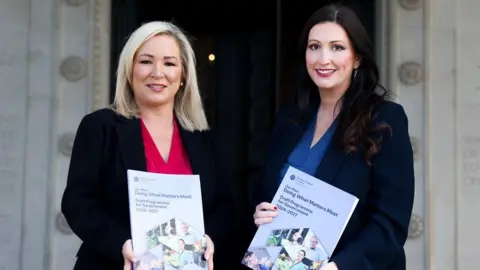 NI Executive Michelle O'Neill with shoulder length blonde hair stands on the left wearing a red top with black blazer. Emma Little-Pengelly with long black hair stands on the right, wearing a dark navy blazer over a blue top. Both women are holding the draft programme document, standing outside Parliament Buildings.