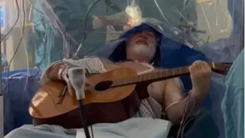 A man with a white beard wearing hospital scrubs with a sheet of blue over his head playing an acoustic guitar