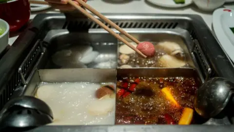 A diner holds a pair of chopsticks over a quad hotpot containing four different types of broth 