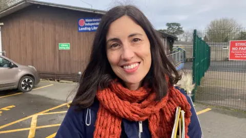 Jenny Kirk/BBC Ava Enrico stands outside the library which is next to a school playground. She wears a navy jacket with a red scarf around her neck.