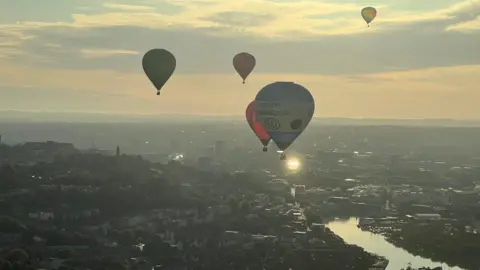 Hot air balloons flying over Bristol. The city and its harbour can be seen below, while the sun rises over the landscape.