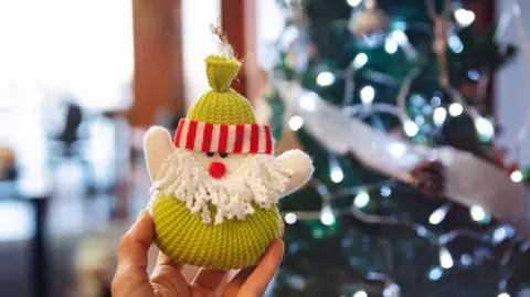 A photo of a person holding a knitted Santa toy in front of a lit up Christmas tree indoors. 