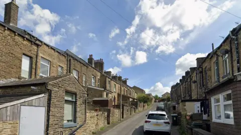 A terraced street on a hill. Houses on either side of the street with cars parked on one side. A blue sky with some clouds above the street.