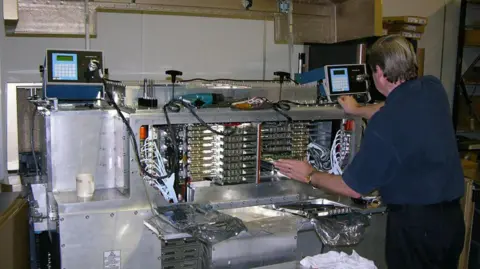 Dan Goldsmith Back of a man in a blue shirt and dark trousers while he looks at one of numerous circuit boards at the back of the big unit, which has been extracted from its box.