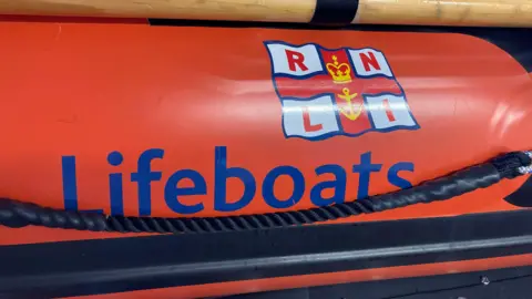 An RNLI flag with the word lifeboats next to it in blue, on an orange background.