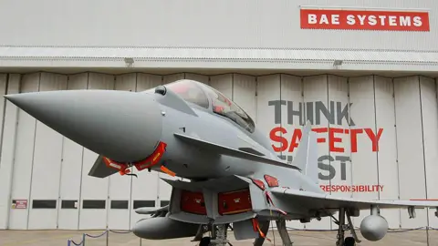 A Eurofighter Typhoon fighter jet at BAE Systems in Warton stands outside a grey warehouse with concertina doors and a red-and-white BAE Systems sign.