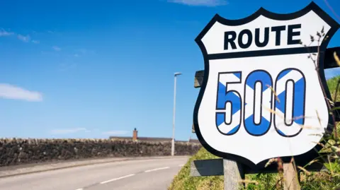 A sign, shaped a bit like a school prefect badge, has been installed by the side of a rural road. The sign is white with the word "route" in black letters and the number 500 in the blue and white colours of Scotland's saltire flag. 
