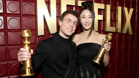 Getty Images Mark Sonnenblick and Ejae pose for a photographer with their Golden Globe trophies. They are smiling and wearing black clothing in front of a maroon backdrop with Netflix written on it in large, gold letters.