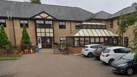 Hallgarth Care Home is a beige-brick two-storey building with a grey roof. It has a small seating area outside and a several car parking spaces. 