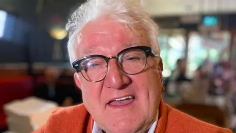 Billy McAneney looking straight at the camera. He is smiling and has white hair and is wearing dark-rimmed glasses. He is wearing an orange jacket with the top of his shoulders visible. The background is blurred.