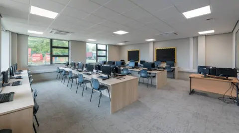 Central Bedfordshire One of the newly developed IT classrooms. There are four rows of wooden desks with black computers and keyboards on top. The carpet in the room is a light grey and the wall and ceiling are white. 