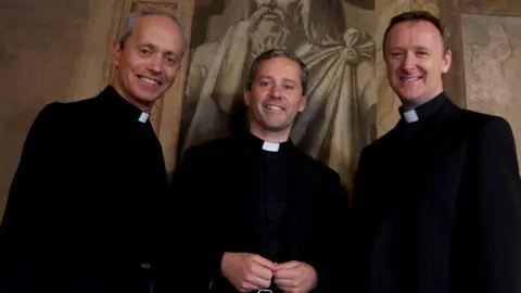 Three priests - Fr Eugene O'Hagan, his brother Fr Martin O'Hagan and Fr David Delargy wearing priest vestments and collars smile at the camera while standing in front of a large statue in the Hotel Colombus in Rome.