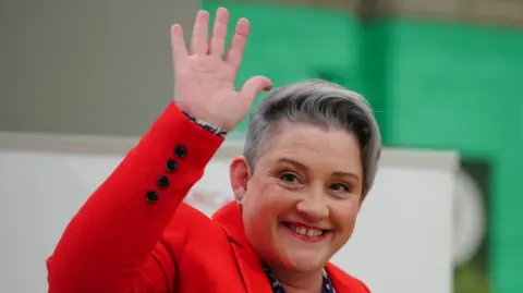 A smiling Ashley Dalton, with short grey hair and a red jacket, waves to the crowd after her election win
