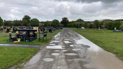 Inside the cemetery, a road down the middle of the fields is dotted with large puddles. There are dozens of gravestones on the left, which are black and grey in colour, and flowers dotted on plots on the right. It is raining.