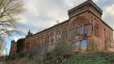 A large sandstone building with arched windows. It is in a derelict state, overgrown with vegetation