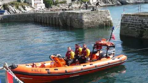 RNLI/ Darren Blight Steve Bennetts at the wheel of an orange inflatable lifeboat, with two other crew. They are in a harbour on a calm day.