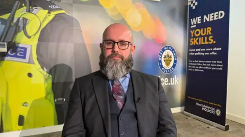 A person wearing glasses sits facing the camera in an indoor interview setting, dressed in a grey suit, white shirt, and patterned tie, with a clip-on microphone attached to the jacket. Behind the person, a large backdrop shows a high-visibility police vest with a radio, alongside the West Midlands Police crest. To the right, a standing banner reads “We need your skills” and promotes careers with West Midlands Police. The scene is brightly lit with a soft, colourful background featuring blurred light effects.
