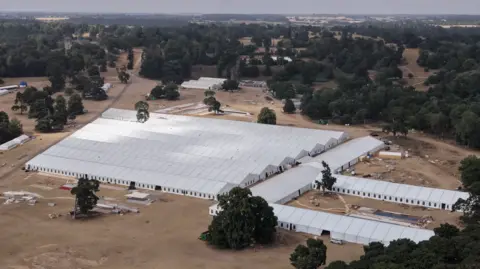 Shaun Whitmore/BBC An aerial photograph showing a massive complex of interconnected white event marquees and tents set up in a large, open field surrounded by dense woodland.