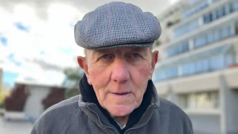 John Mahoney in a fleece jacket and a blue and red patterned flat cap. 